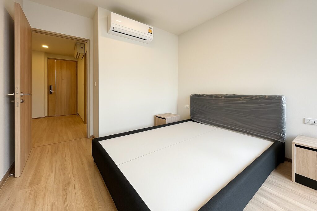 Modern minimalist bedroom interior at XT Phayathai condominium in Bangkok, featuring a queen-sized bed frame with upholstered headboard, light wood flooring, and air conditioning, ideal for urban living.